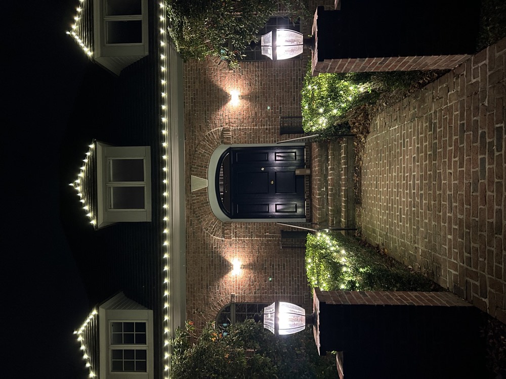 A brick house with lit dormers and hedges lining the brick walk to the front door